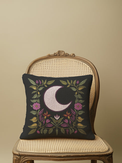 Black woven throw pillow with an embroidered crescent moon and floral pattern in soft ivory, green, purple, and gold, featuring a textured finish.